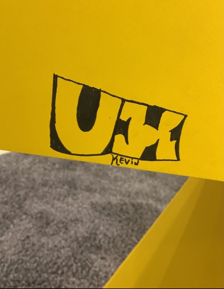 “UX” desenhado à mão em caneta preta numa superfície amarela, assinado Kevin